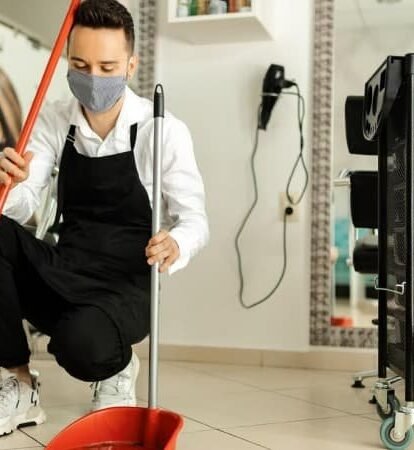 Reinigungskraft kehrt Boden mit Besen und Schaufel in modernem Salon für hygienische Gewerberäume.